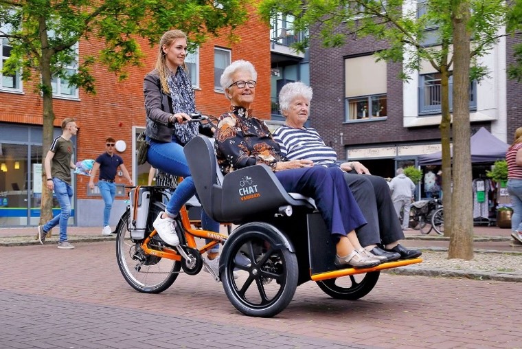 Foto einer Rikscha, es zeigt eine Frau, die das Fahrrad fährt, und zwei ältere Frauen, die vorne sitzen und gefahren werden.